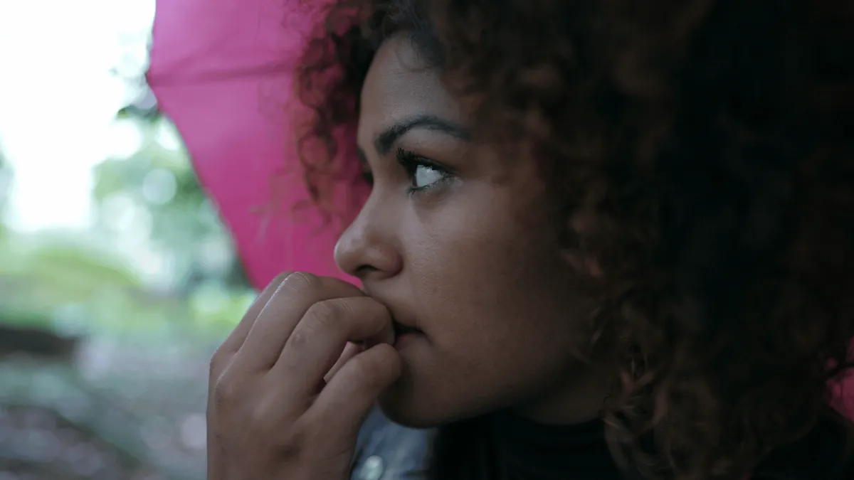 Beautiful black woman biting her nails, worried or anxious and deep in thought, outside in the rain, standing with a pink umbrella.