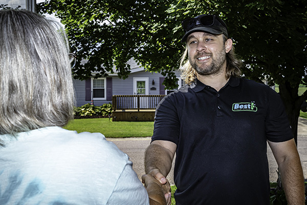 Professional carpet cleaner from Best Steam Clean greeting a homeowner with a handshake in a residential driveway, wearing company polo shirt with house visible in background