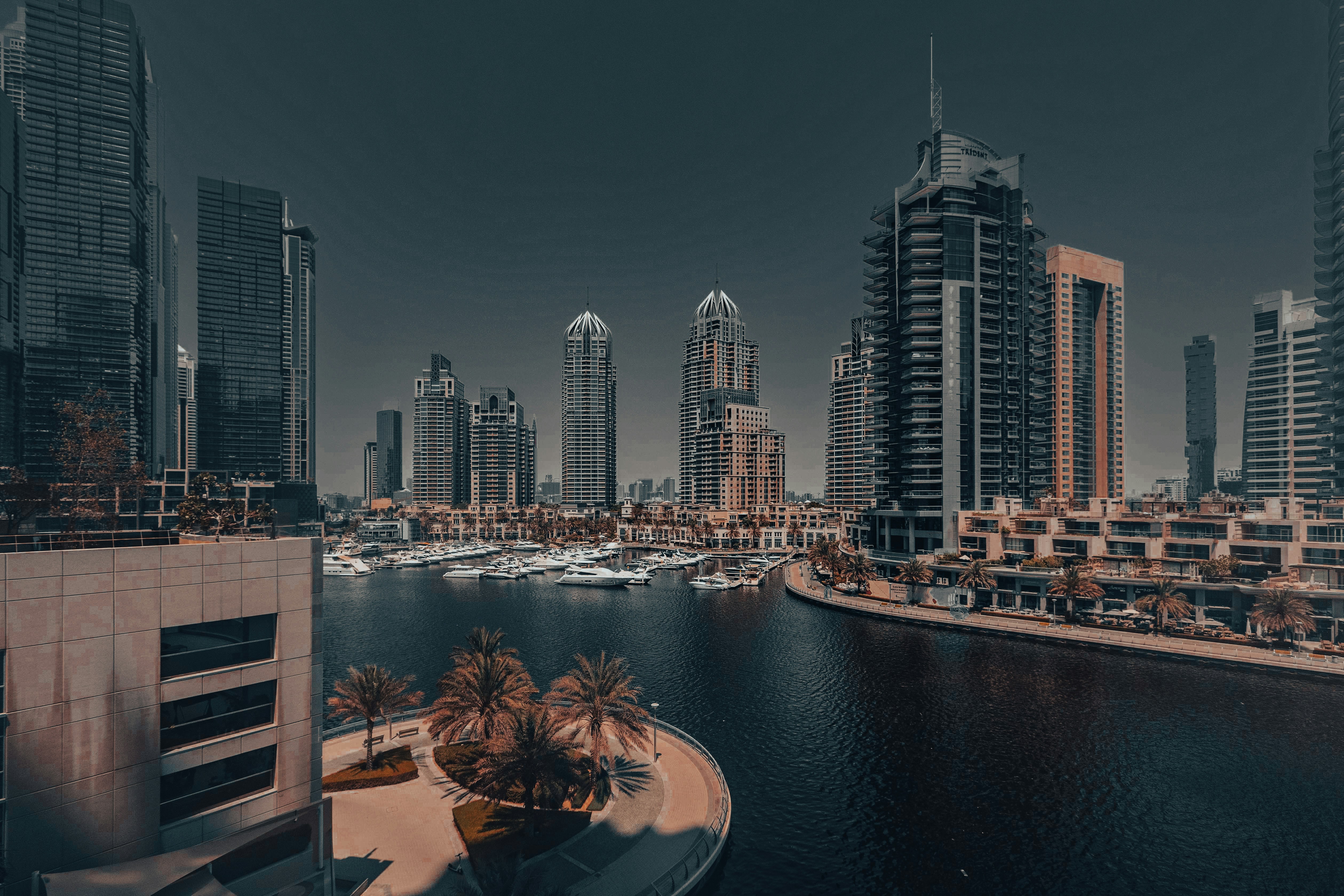 “Modern waterfront city skyline with luxury yachts, high-rise buildings, and palm trees under a dramatic sky, showcasing a vibrant urban marina destination – Alora Travels”