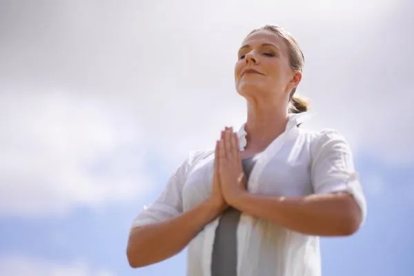 A serene middle-aged woman stands outdoors against a bright, cloudy sky. She is dressed in a light blouse with her hands in a prayer position at her chest, eyes closed, and a calm, peaceful expression on her face—radiating a sense of mindfulness and inner strength.