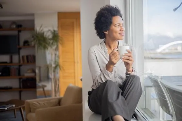 Smiling midlife woman enjoying a warm drink by the window, representing relaxation and natural ways to support digestion and reduce bloating.