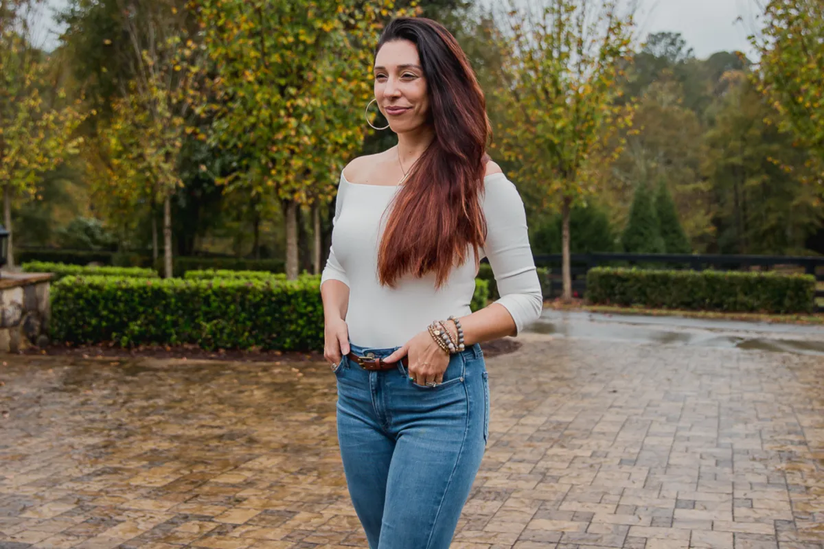 CeCe standing in front of a garden wearing a grey off-the-shoulder top