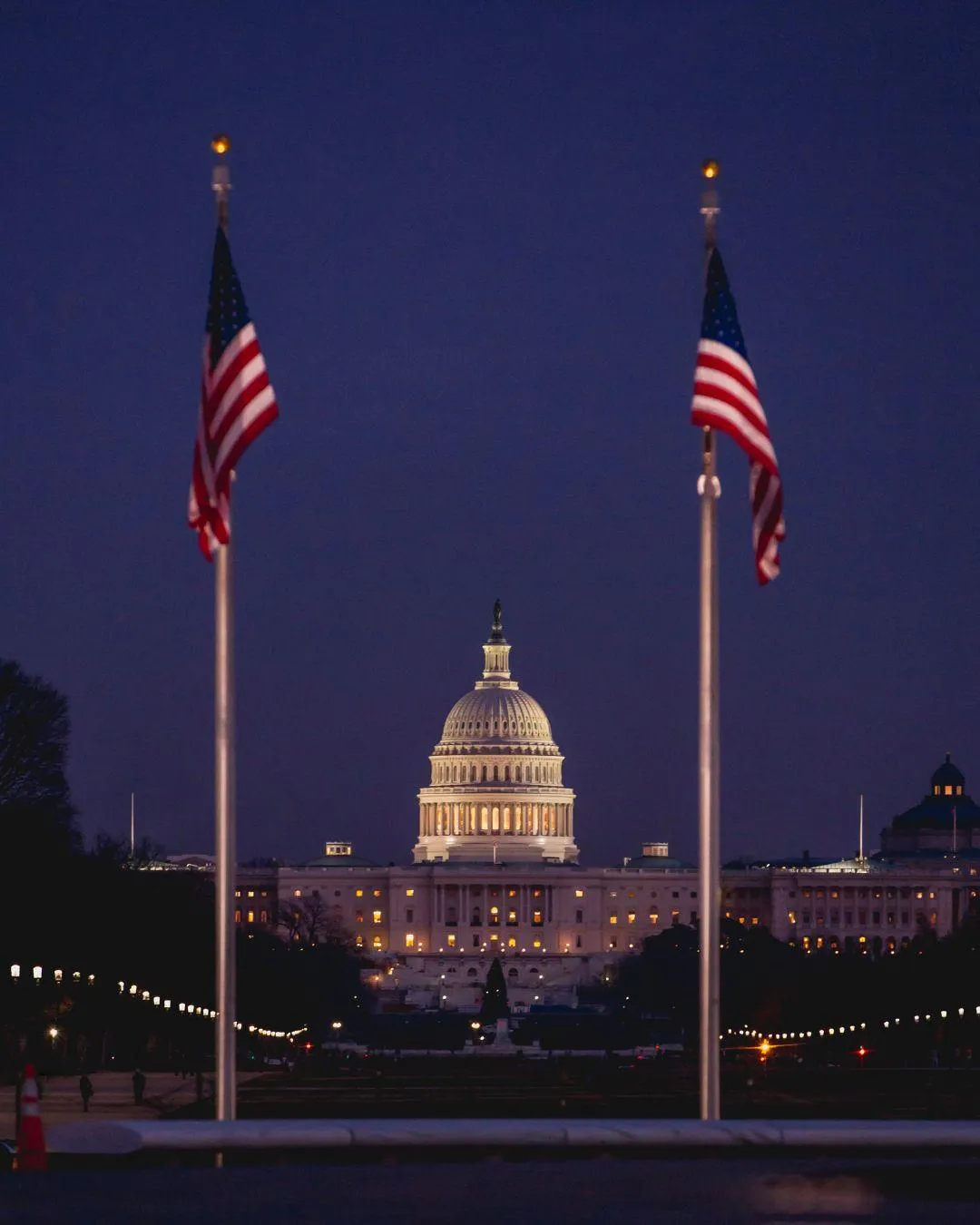 The U.S. Capitol Building is lit up at dusk, seen between two American flags flying on flagpoles.