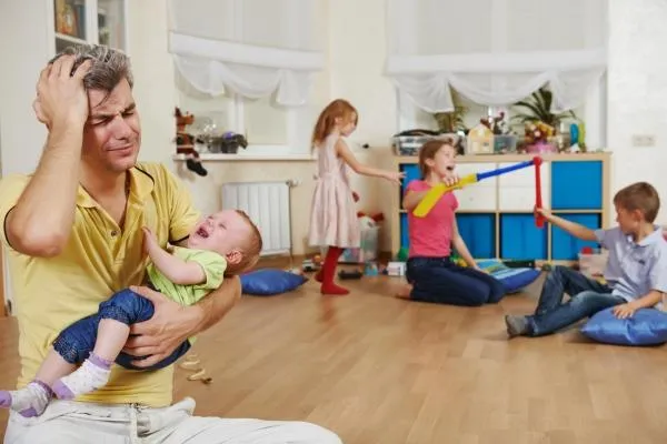man looking distressed while carrying a crying baby and other kids playing near him - fear of parenting failure