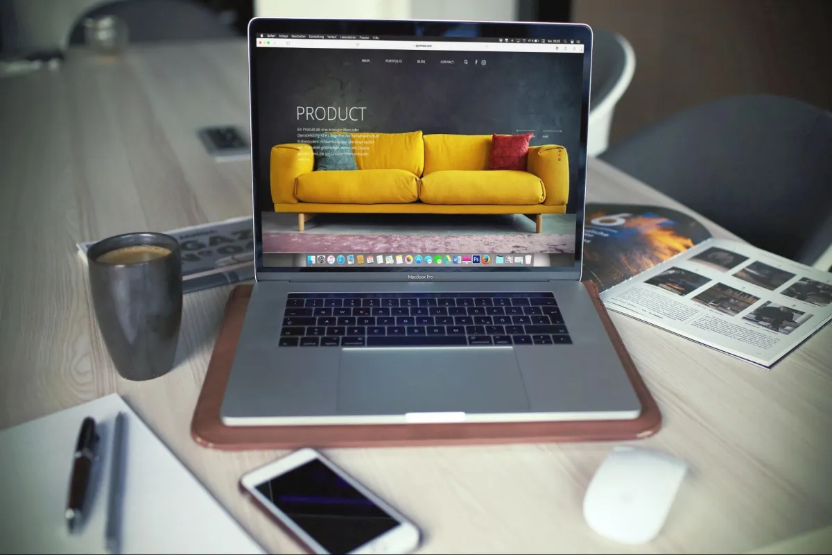 Laptop displaying a modern product website with a yellow sofa on a wooden desk, alongside a coffee cup, smartphone, and magazine.