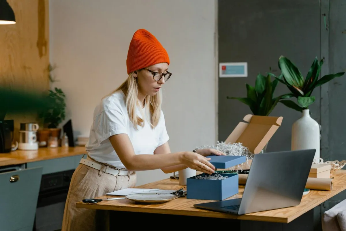 Smiling woman holding a gift box and using a laptop at a table, suggesting online shopping or gift exchange.