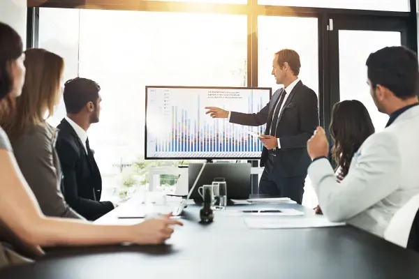 People seated at a conference table with a man standing in front of a board and presenting
