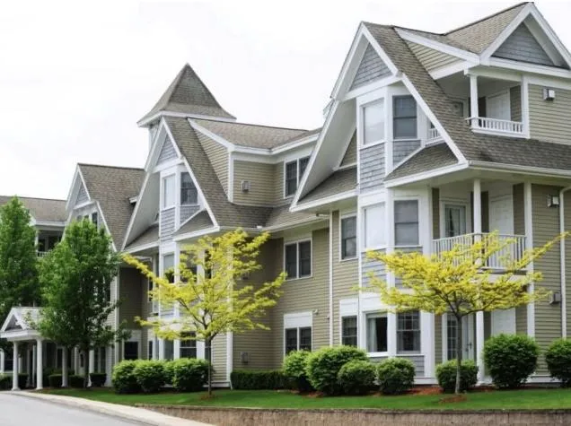 Row of beige multi-story residential buildings with white trim, peaked roofs, and neatly landscaped greenery in front.