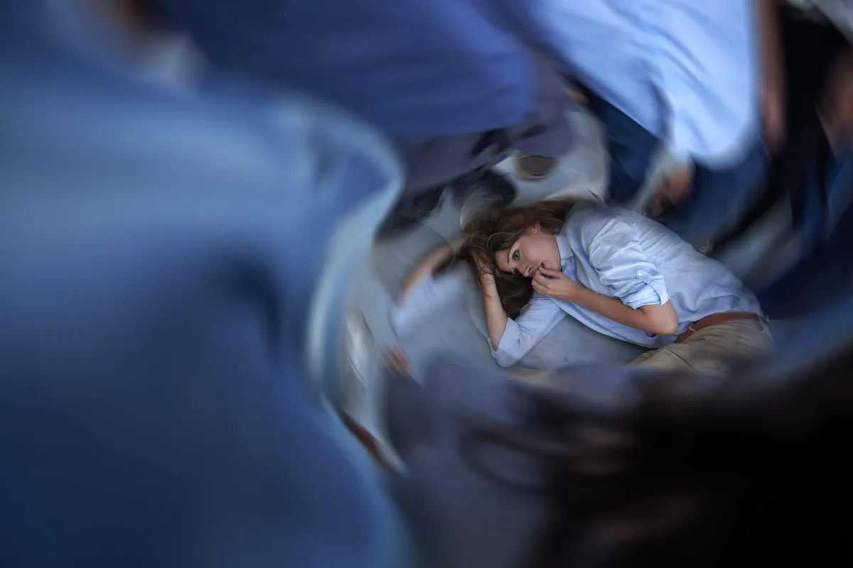 A woman lies curled on the floor in a fetal position, overwhelmed and anxious, surrounded by blurred figures rushing past her. The image conveys sensory overload and emotional distress, with a swirling effect representing overstimulation and internal chaos.