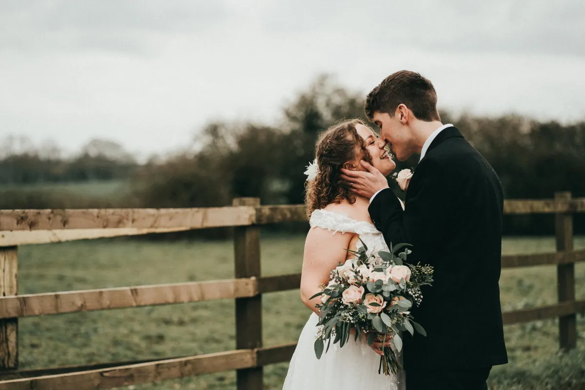 Photo of wedding couple standing in front of a fence and leaning in for a gentle kiss at Milling Barn Wedding Venue