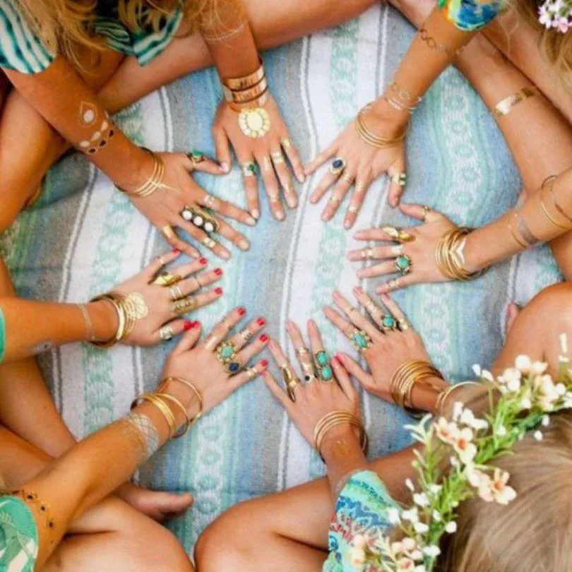 A group of people sitting in a circle, touching hands at the center with various rings and bracelets, over a patterned textile, symbolizing unity and friendship.