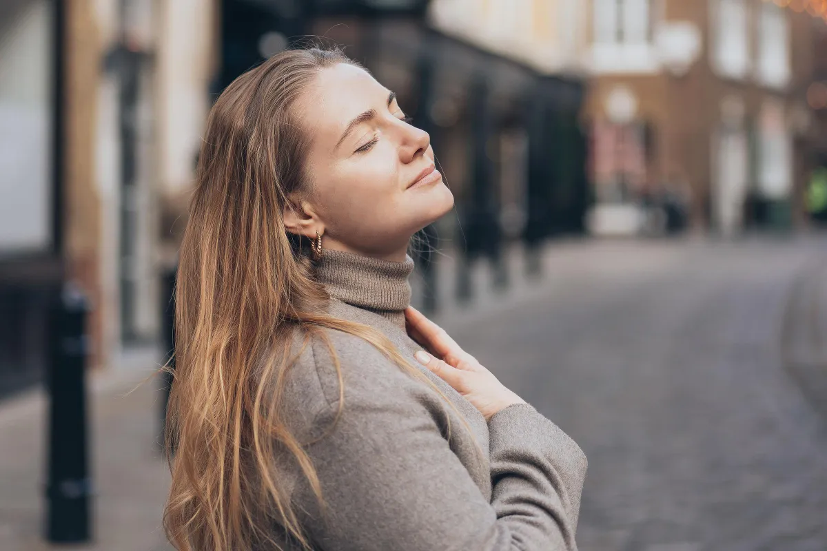 A serene woman with long blonde hair closes her eyes and gently touches her chest while standing on a quiet street. She appears content and peaceful, wearing a beige turtleneck sweater.