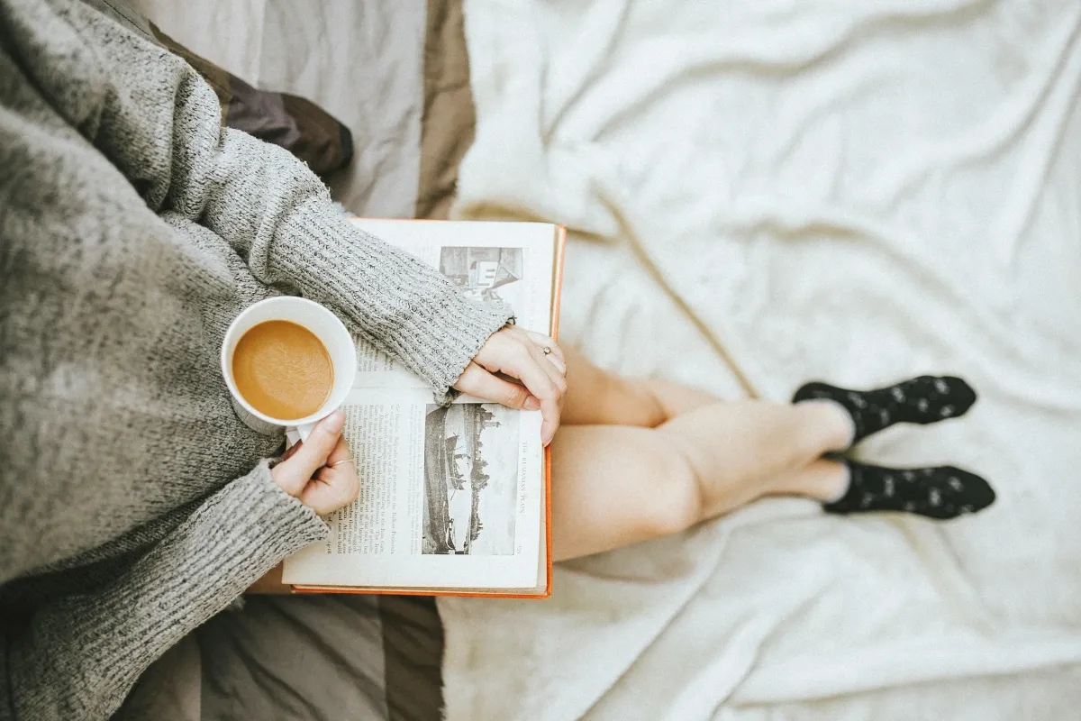 A person sitting on a cozy bed with a soft gray sweater, holding a cup of coffee in one hand and a book in the other, creating a calm and relaxed atmosphere.