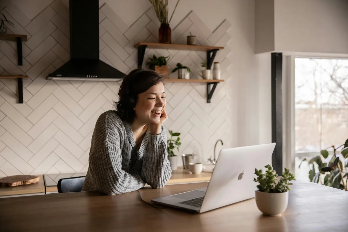 Smiling woman in a cozy kitchen setting, wearing headphones and working on a laptop, symbolizing intentional living and online personal growth.