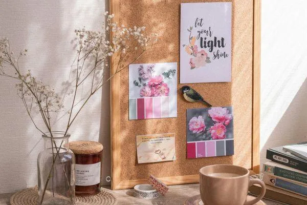 A rustic desk setup with a cork board holding cards, including one that reads 'Let your light shine.' A journal, a coffee cup, and a vase with dried flowers are also on the desk, bathed in warm sunlight.
