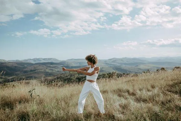 A person with curly hair in white clothing performs a dynamic, expansive movement pose in a wide, grassy field with rolling hills and a cloudy sky in the background.