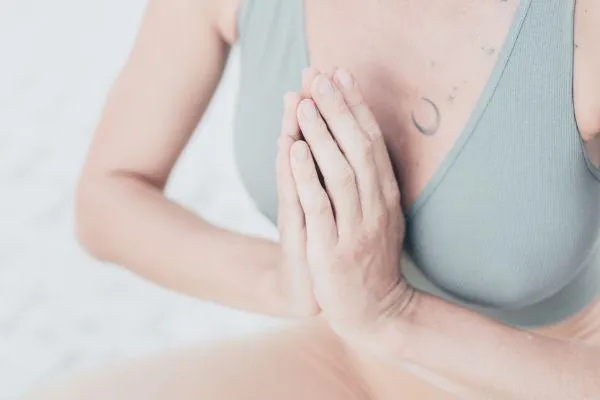 Close-up of a person's hands pressed together in a prayer or anjali mudra position, resting at chest level. They are wearing a light-colored sports bra or top, and a subtle moon and stars tattoo is visible on their upper chest. The background is soft and out of focus.