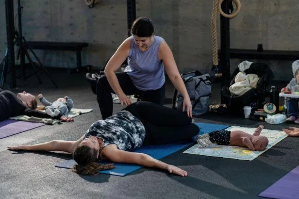 A new mom and her baby perform a squat in a postpartum rehab fitness class. The Stittsville gym is open and inviting, and other moms can be seen with their babies exercising in the background.
