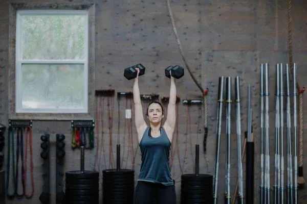 A woman does a back squat using a barbell with weights during a personal training session. Another member of the gym can be seen behind her working out using different equipment. The setting is a clean Stittsville gym with various weights, barbells, and other exercise equipment lining the walls.