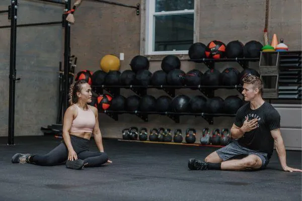 A Between The Bumpers Stittsville Physiotherapist and his client are seated on the gym floor mid-stretch, having a focused conversation about the benefits of the strtch for her rehabilitation from injury, with medicine balls and kettlebells in the background.