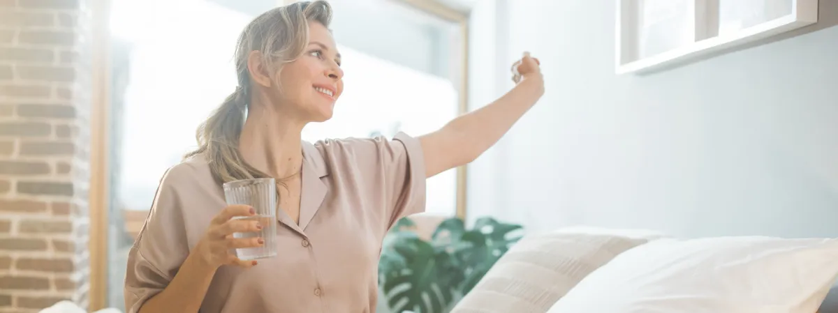 Smiling woman stretching her arm while holding a glass of water in a bright and cozy bedroom, symbolizing relief and vitality, potentially achieved through Hormone Replacement Therapy (HRT) for managing menopause symptoms.