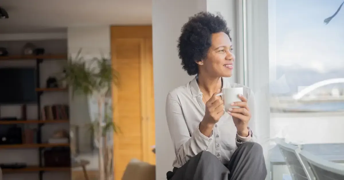 Smiling woman sitting by a bright window holding a coffee mug, looking outside peacefully.