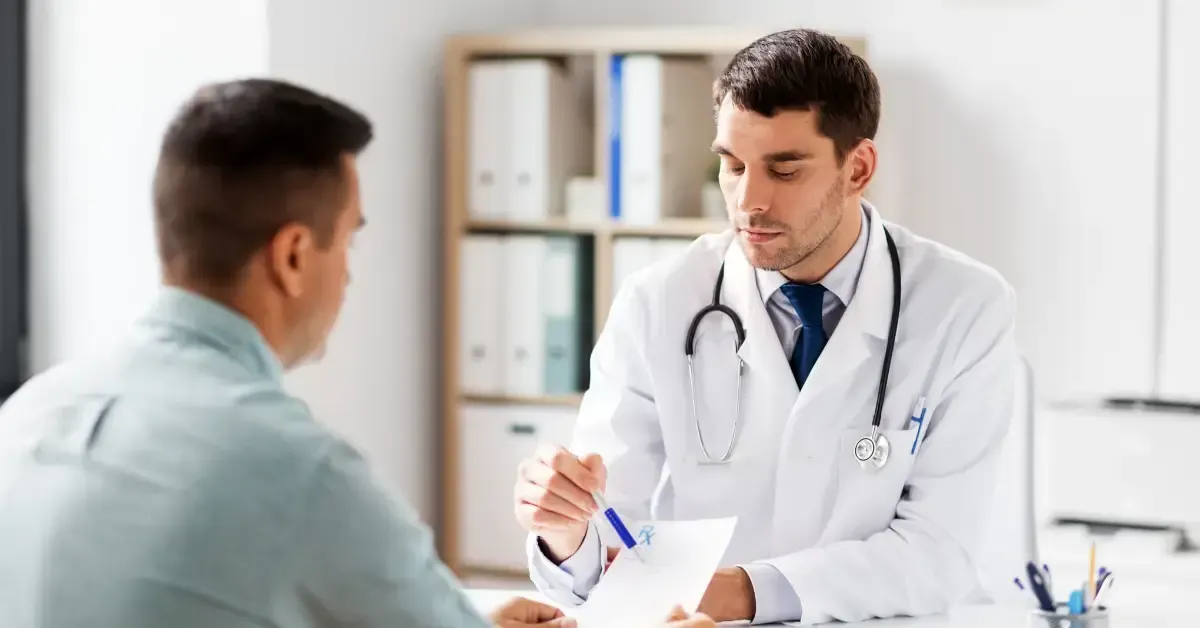 A doctor in a white coat with a stethoscope talks to a male patient across the desk while reviewing paperwork.