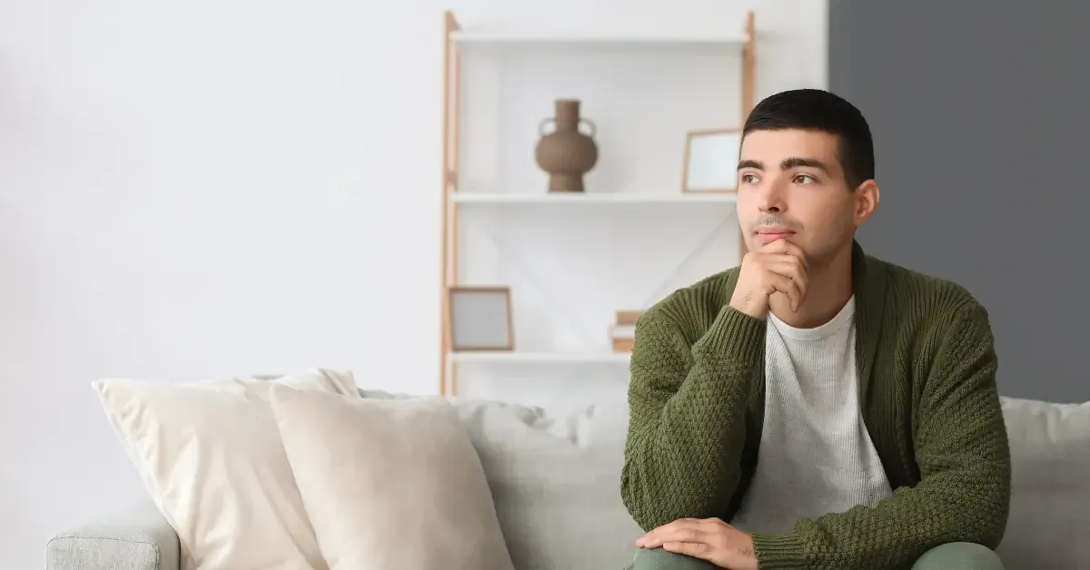 A young man sits on a sofa resting his chin on his hand, looking thoughtful and pensive.