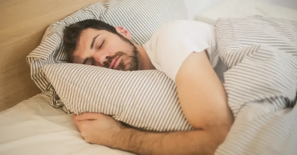 A man with a beard peacefully sleeping on his side in bed, covered with a striped blanket.