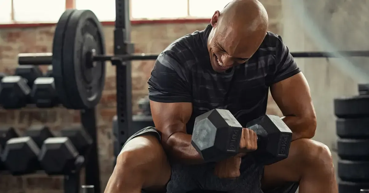 A muscular man intensely lifting a heavy dumbbell while seated in a gym, surrounded by weightlifting equipment.