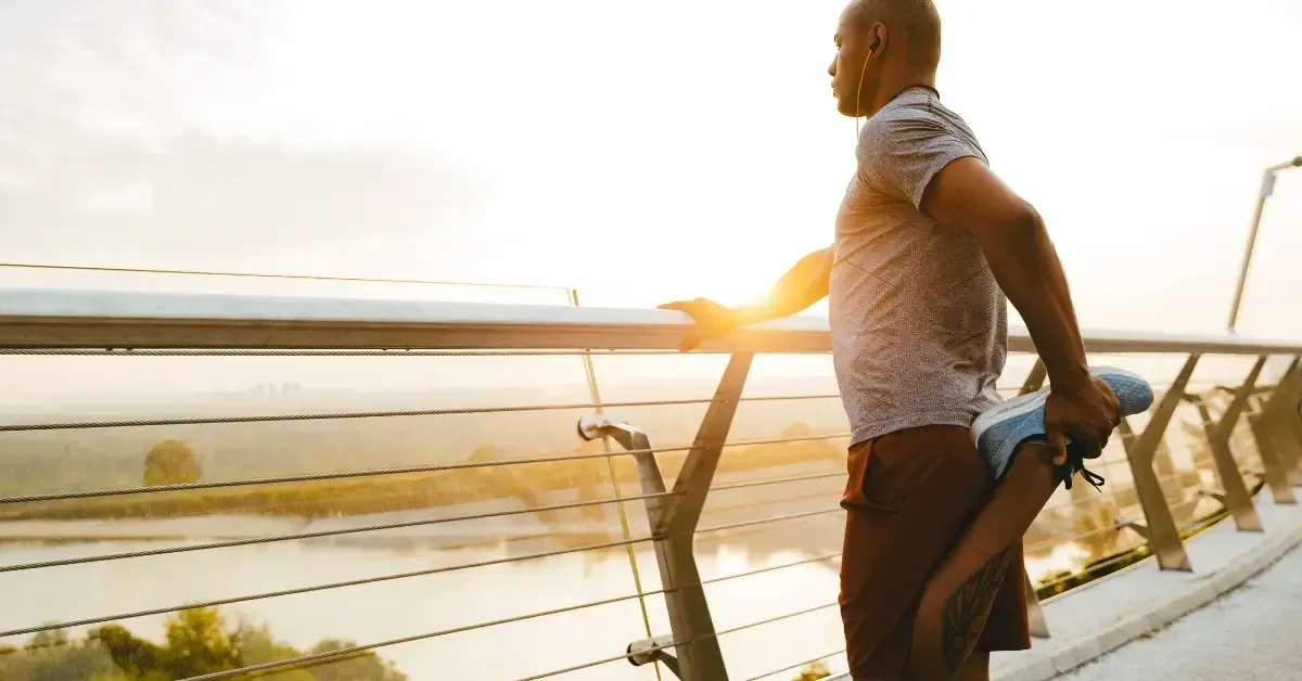 Man stretching his leg while holding onto a railing outdoors at sunrise, preparing for a workout.
