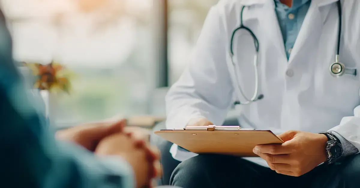 A doctor in a white coat holds a clipboard while sitting across from a patient, whose hands are clasped in the foreground, during a consultation in a softly lit medical office.