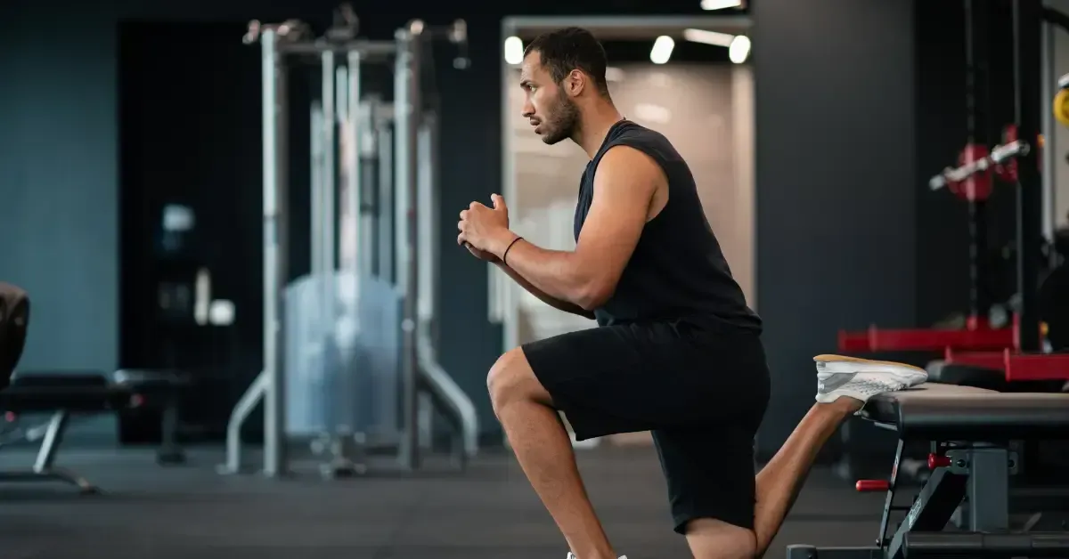 A man in athletic clothing performs a lunge stretch in a gym, with one foot resting on a bench and exercise equipment visible in the background.