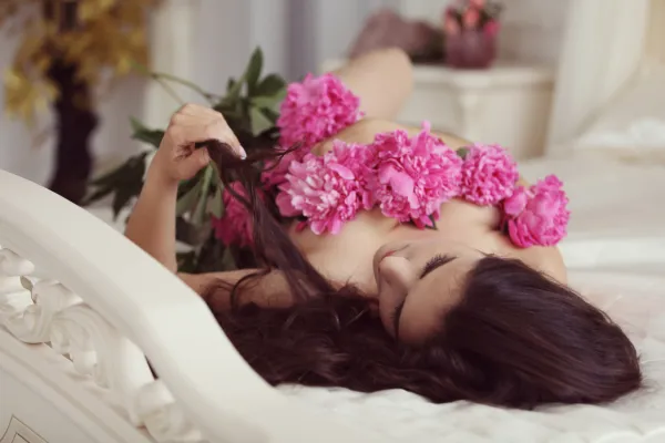 Dark haired caucasian woman laying naked on a white bed with a bunch of pink chrysanthemum flowers draped over her chest