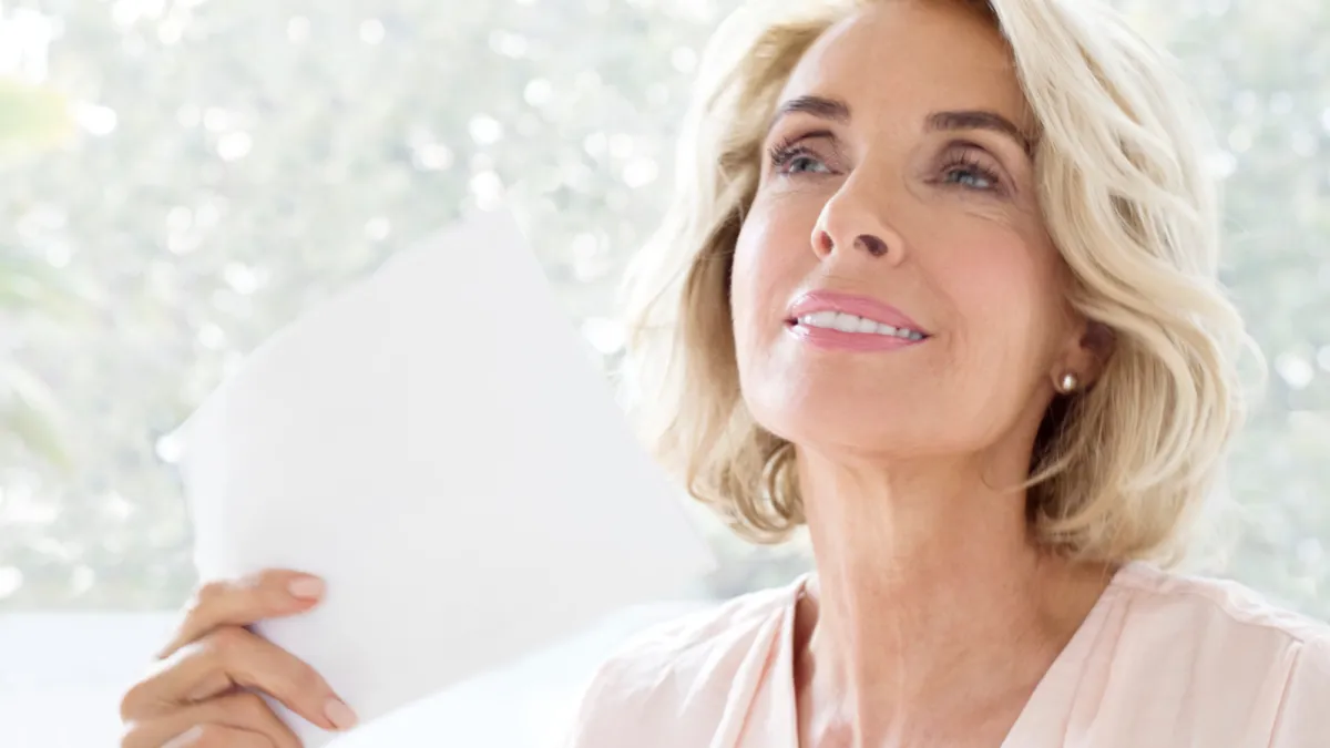 Head and shoulders image of a mature caucasian woman with blonde, shoulder length hair and wearing a pale pink top using a piece of white paper to fan herself. She has a slight smile on her face and is looking slightly off camera.