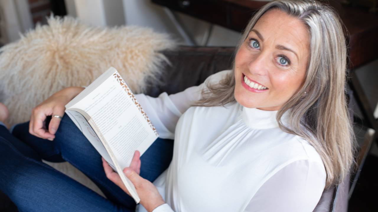 Blonde haired Caucasian woman with blue eyes sitting across a chair in a white bluse and jeans holding a book