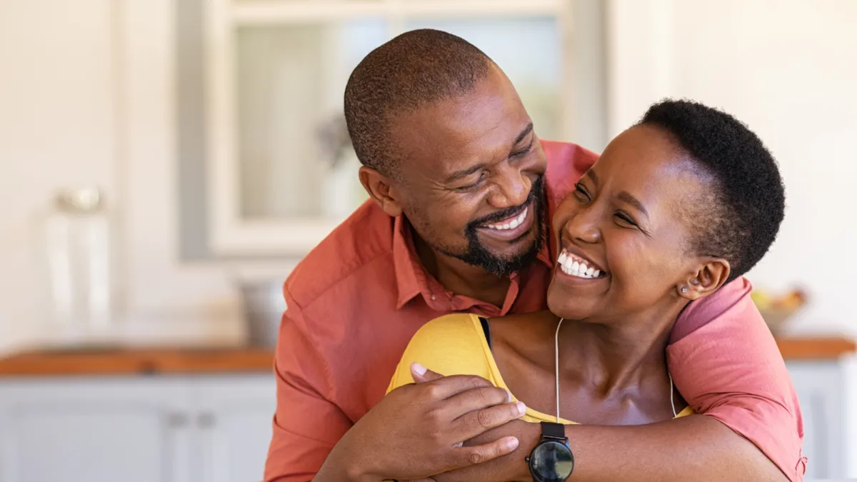 Black woman in a yellow top being hugged from behind by a black man in a red shirt. Both are smiling and very happy