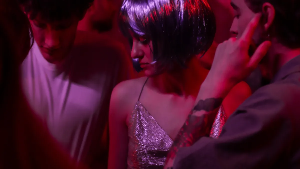 Woman in a purple bobbed wig wearing a sparkling strapped dress with bare arms and shoulders, looking to her right. Dark haired caucasian male in white tshirt looking down to her right and to her left a dark-haired man with his finger in his ear. Club lighting, pinks, purples and reds.