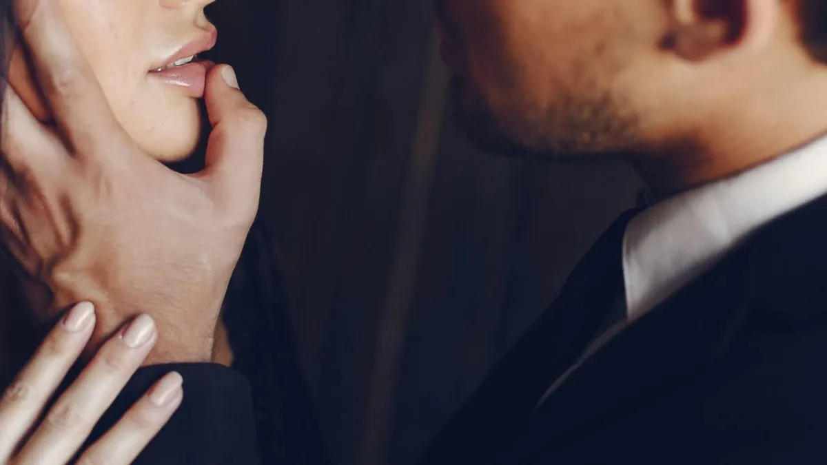 Man in dark suit and tie with white shirt and stubble placing his hand on the face of a caucasionwoman, his thumb on her lips, wearing lipgloss, her hand places on his wrist seductively