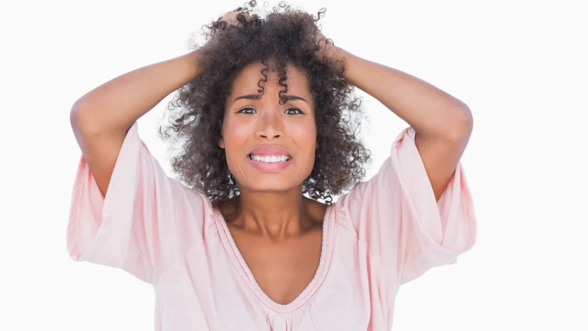 Woman of colour with her hands in her hair wearing a pale pink baggy, v-neck t-shirt, looking worried or anxious