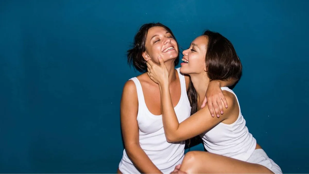 Two caucasian women with brown bobbed hair, both wearing white vests and shorts smiling and laughing as they embrace