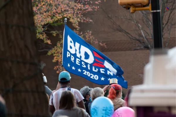 Crowd of people at a political rally holding a 'Biden President 2020' flag, with autumn trees and city buildings in the background.