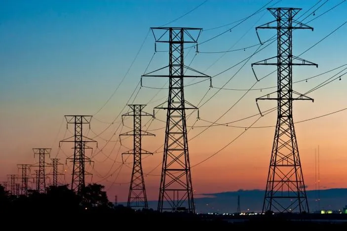 Silhouettes of tall power transmission towers against a vibrant sunset sky, with power lines stretching across the horizon, set against a distant city glow.