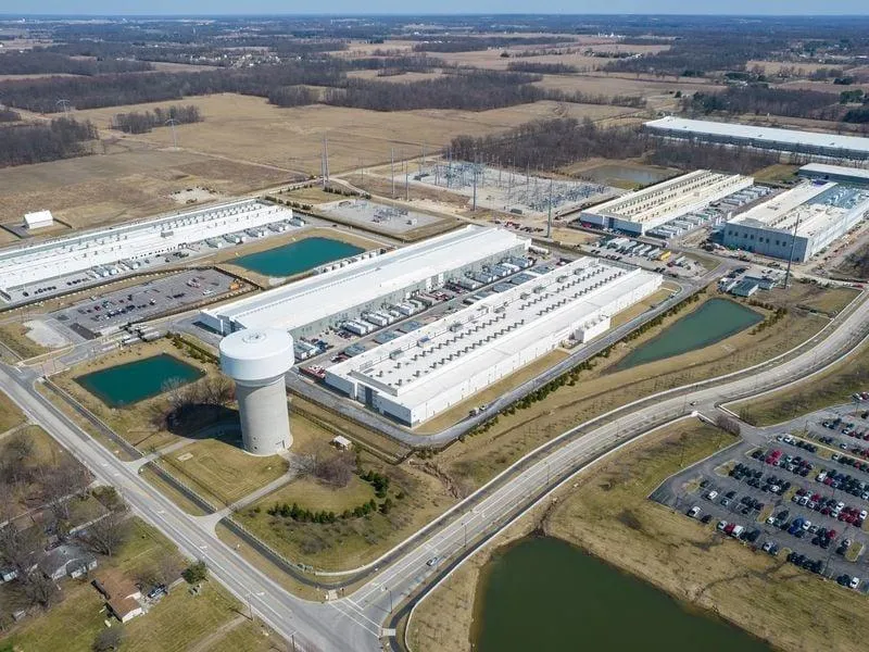 erial view of a sprawling industrial facility featuring long white buildings, retention ponds, a prominent water tower, parking areas filled with vehicles, and surrounding open fields under a clear sky.