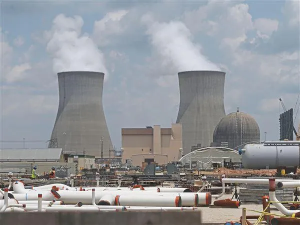 Nuclear power plant with two large cooling towers releasing steam, a dome-shaped reactor building, industrial buildings, pipes, and equipment in the foreground under a partly cloudy sky.