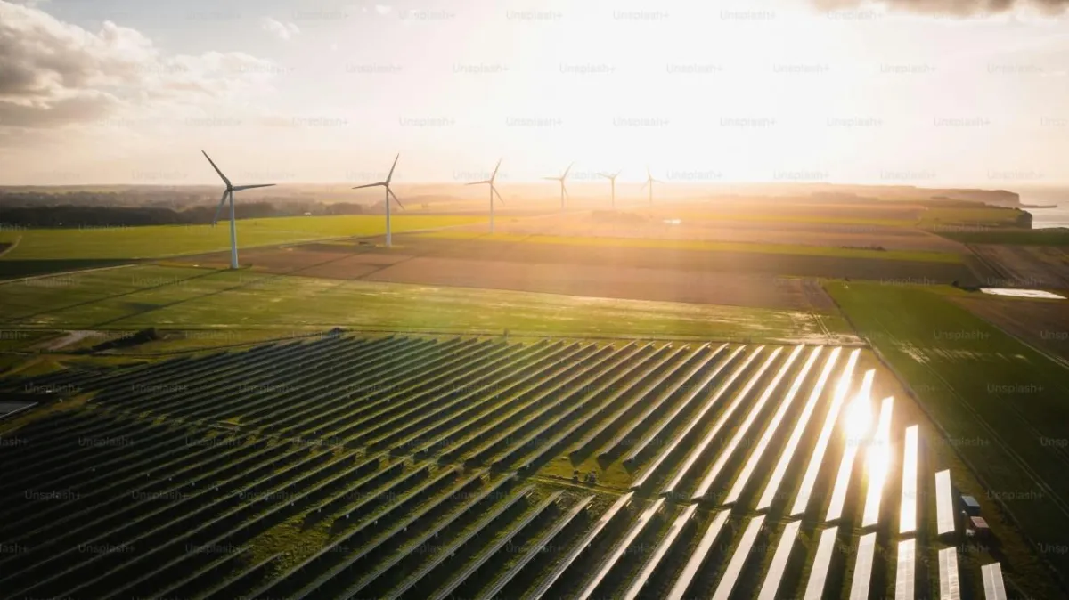 Aerial view of a expansive solar panel farm in the foreground with rows of panels on green fields, transitioning to wind turbines in the background under a golden sunrise or sunset sky with scattered clouds and distant landscape.