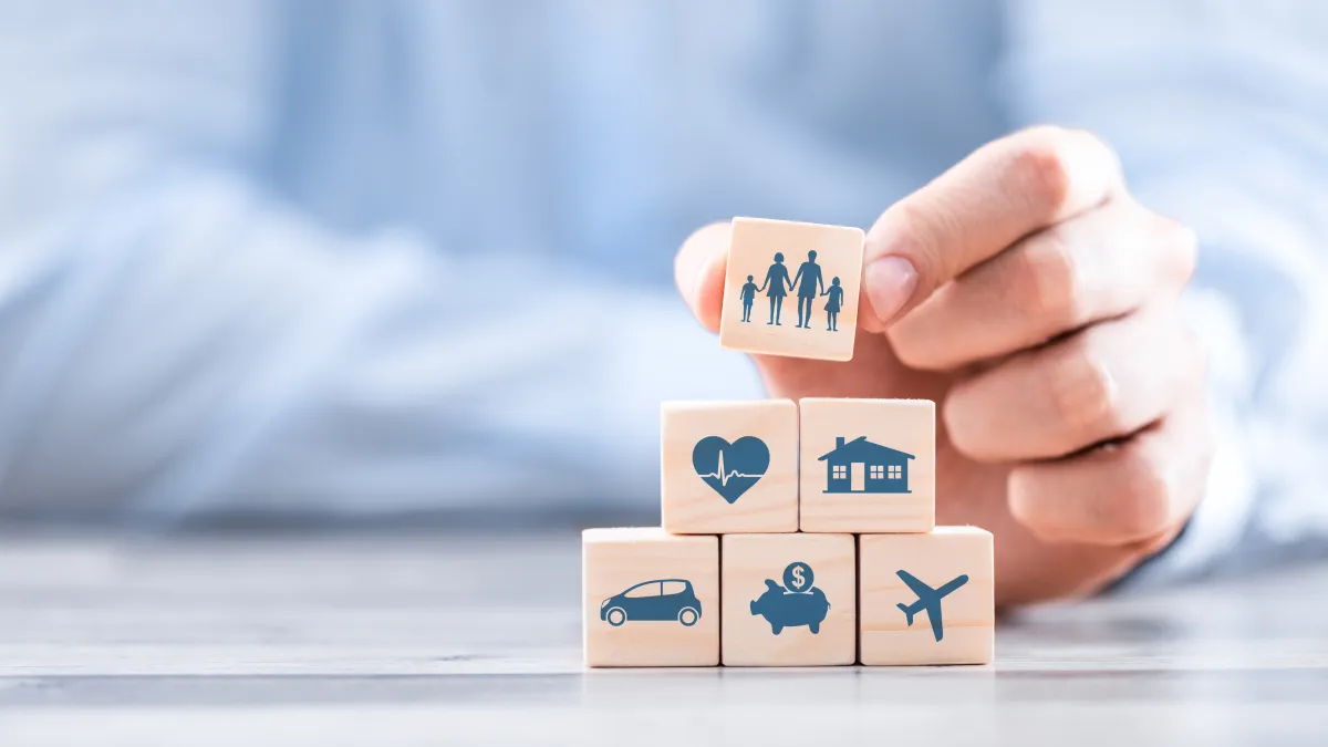 Hand stacking wooden blocks with family, heart, and house icons, symbolizing life, health, and home insurance protection.