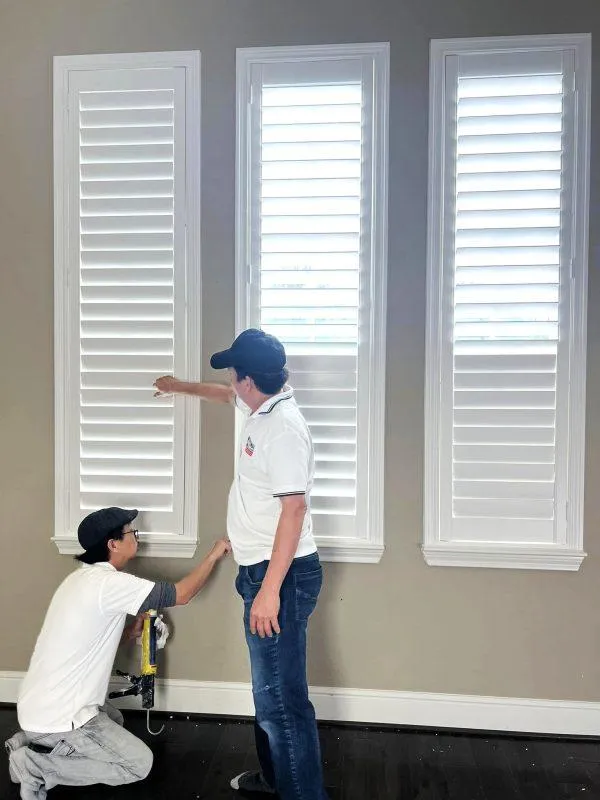 A professional installer in uniform carefully fitting a white full height shutter into a window frame.