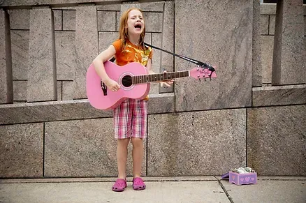A red-headed girl in a colorful outfit plays a pink guitar while singing along.
