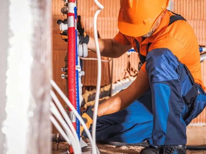  A plumber in an orange hard hat and blue overalls crouches, working on a system of red and blue plastic pipes connected to a wall